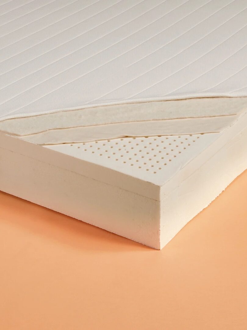 Cross-section of a mattress showing multiple layers of foam, including a perforated layer, against a peach-colored background.