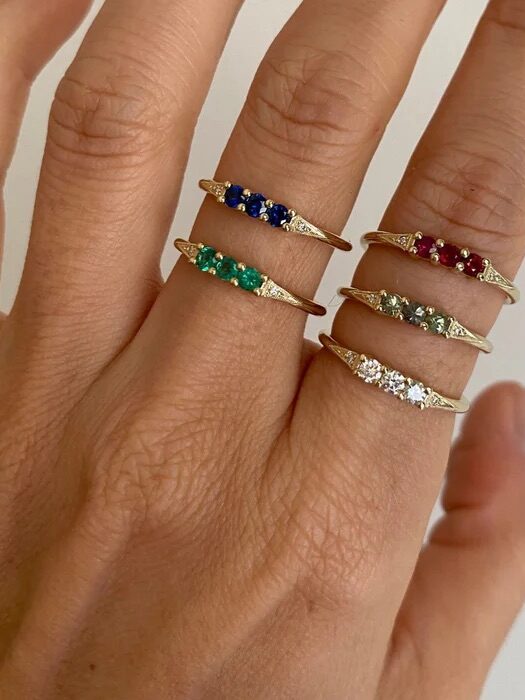 A hand wearing three gold rings, each set with different colored gemstones: blue, red, green, and clear. The rings are worn on the index, middle, and ring fingers.