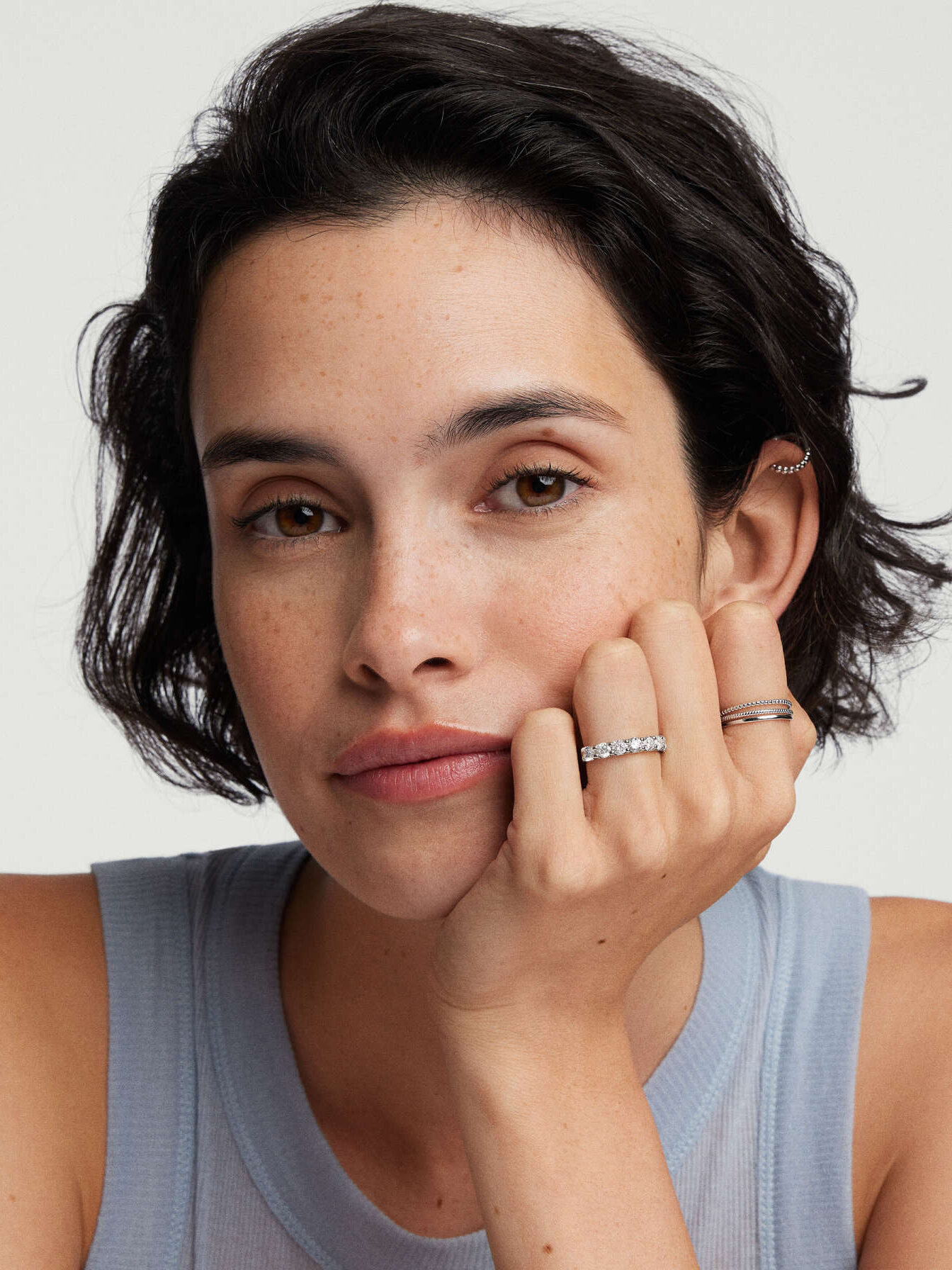 A person with short hair rests their head on their hand, wearing a sleeveless top, a ring on their index finger, and an ear cuff.