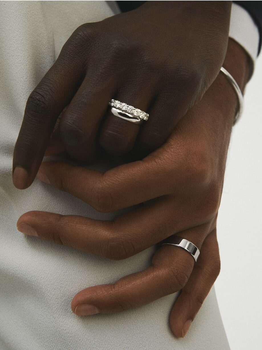 Close-up of two hands, one wearing a silver ring with stones and the other wearing a plain silver band. Both are intertwined against a white and black background.