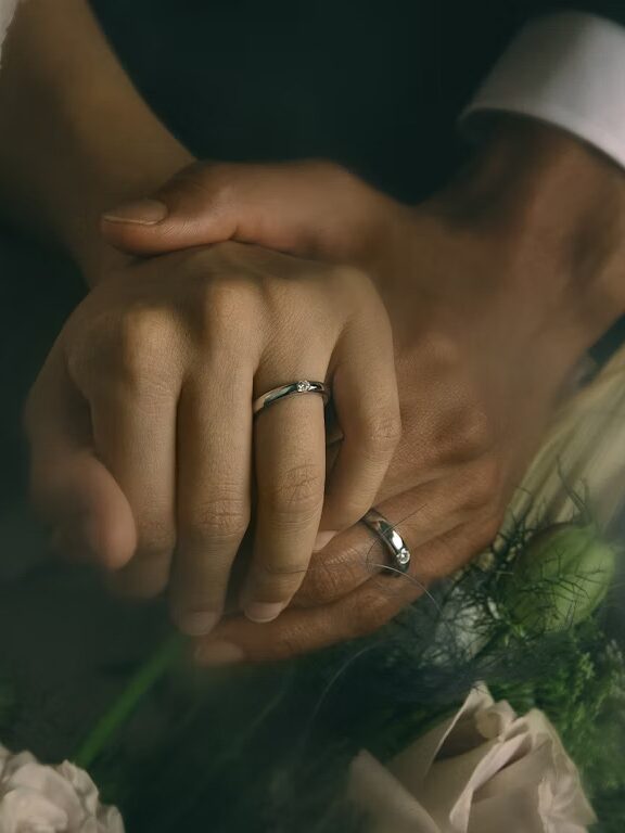 Two hands, each wearing a wedding ring, are clasped together against a background of white flowers and greenery.