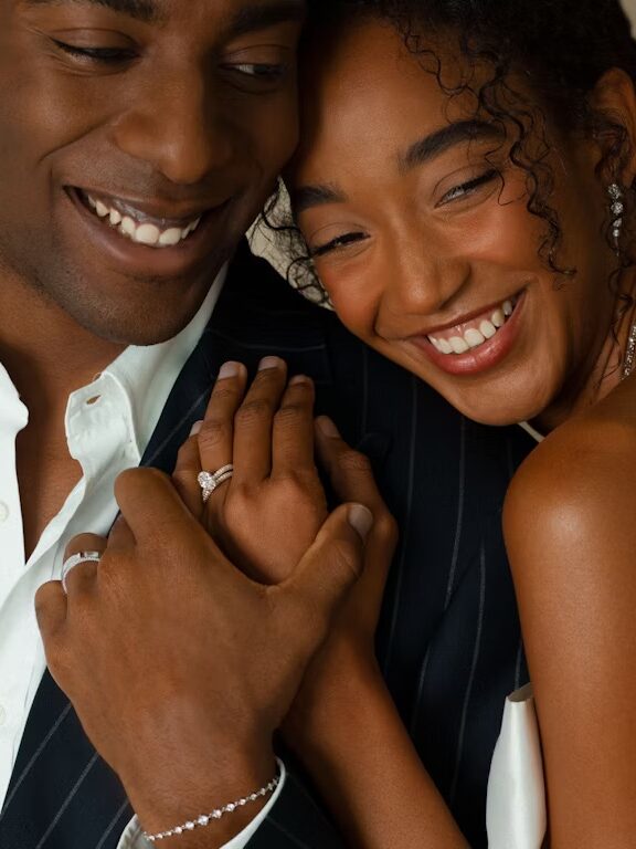 A smiling couple, dressed formally. The woman, wearing a white dress, holds her hand with an engagement ring over the man's chest, both showing affection. The man is in a dark pinstripe suit.