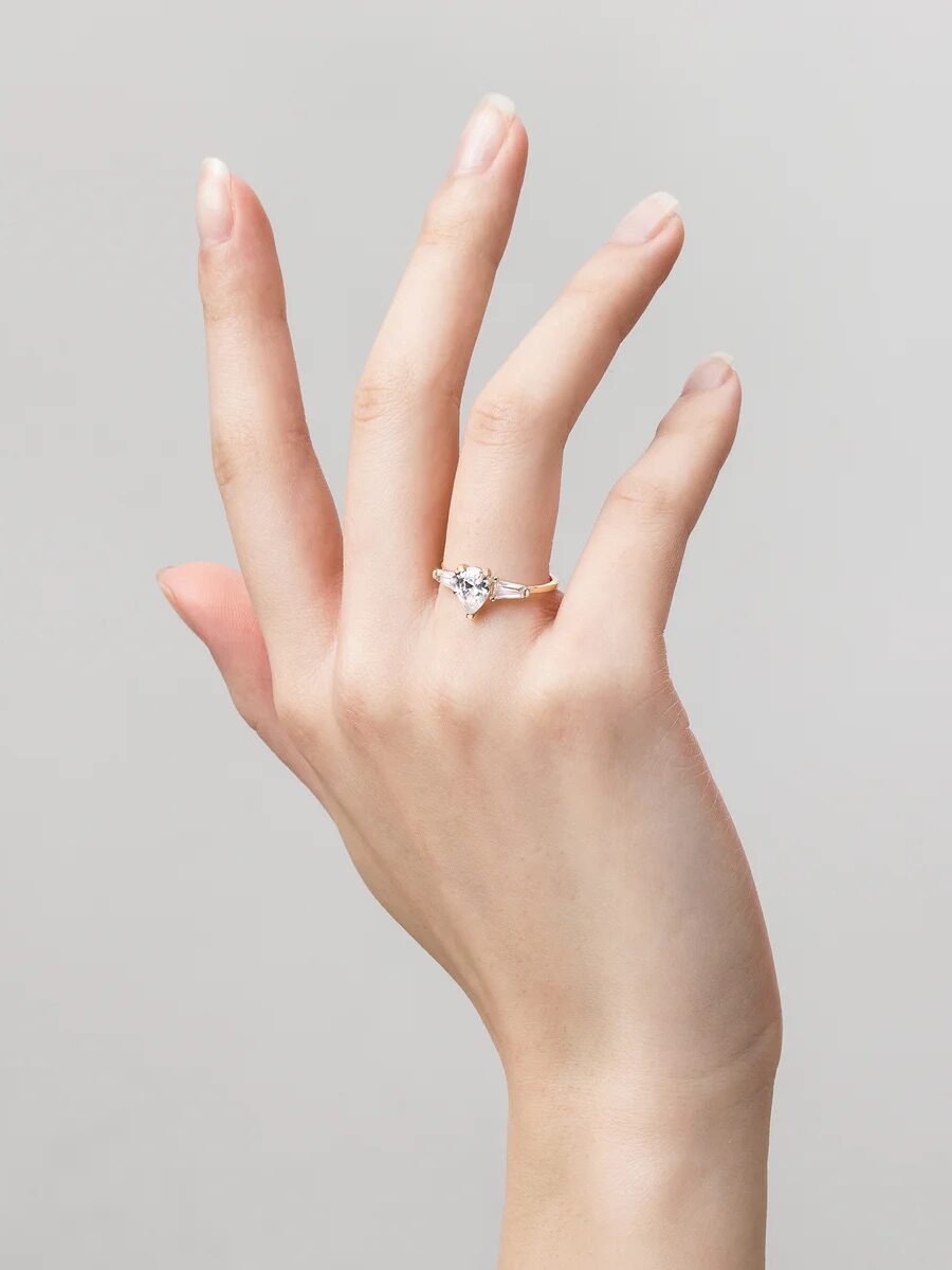 A hand with a diamond ring on the ring finger is shown against a gray background.