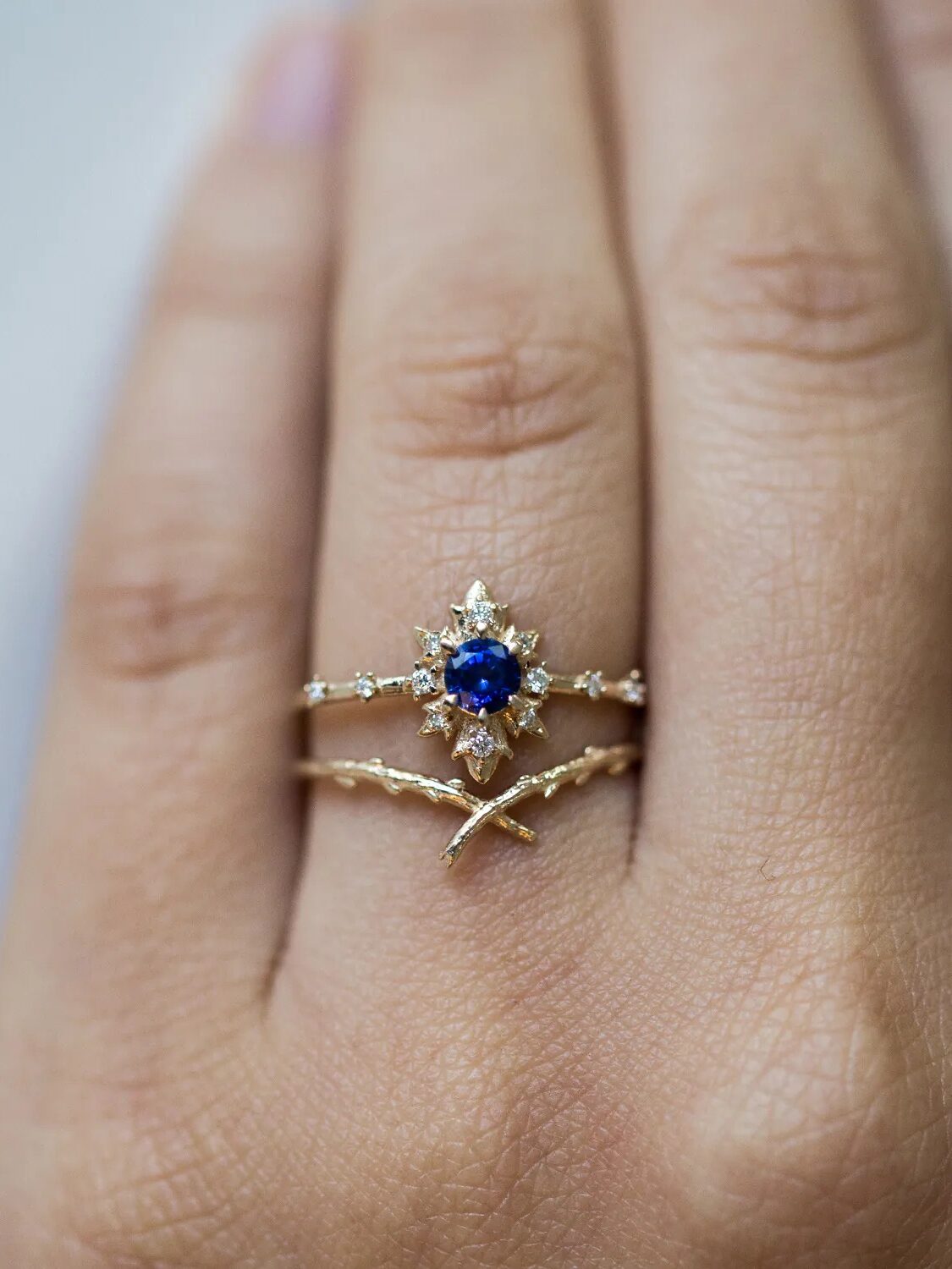 Two rings are worn on a hand: a gold ring with a blue gemstone surrounded by small clear stones, and a second gold ring with a twisted design.