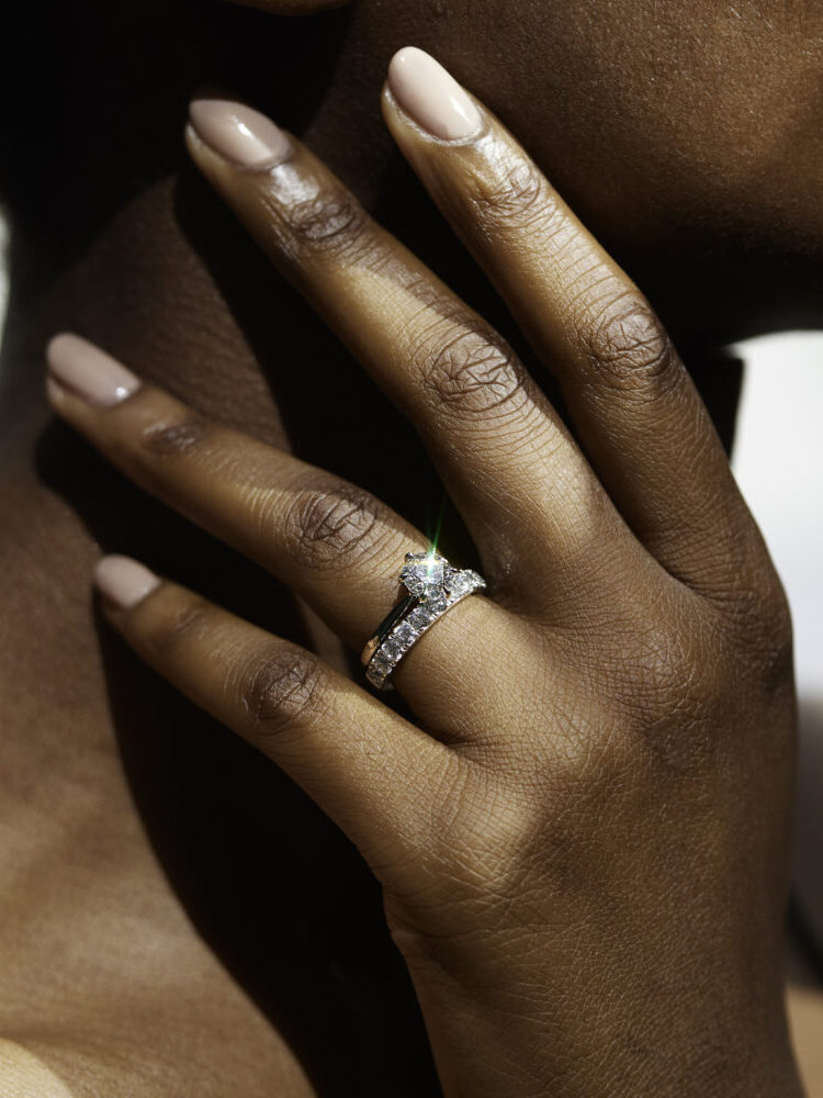 A person with manicured nails touches their neck, showcasing a sparkling diamond ring on their finger.