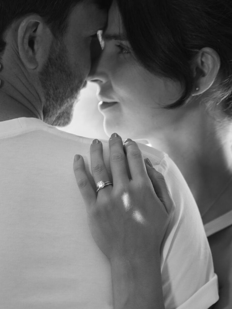 A close-up black and white image of a couple embracing with the woman's hand resting on the man's shoulder, showcasing an engagement ring on her finger.