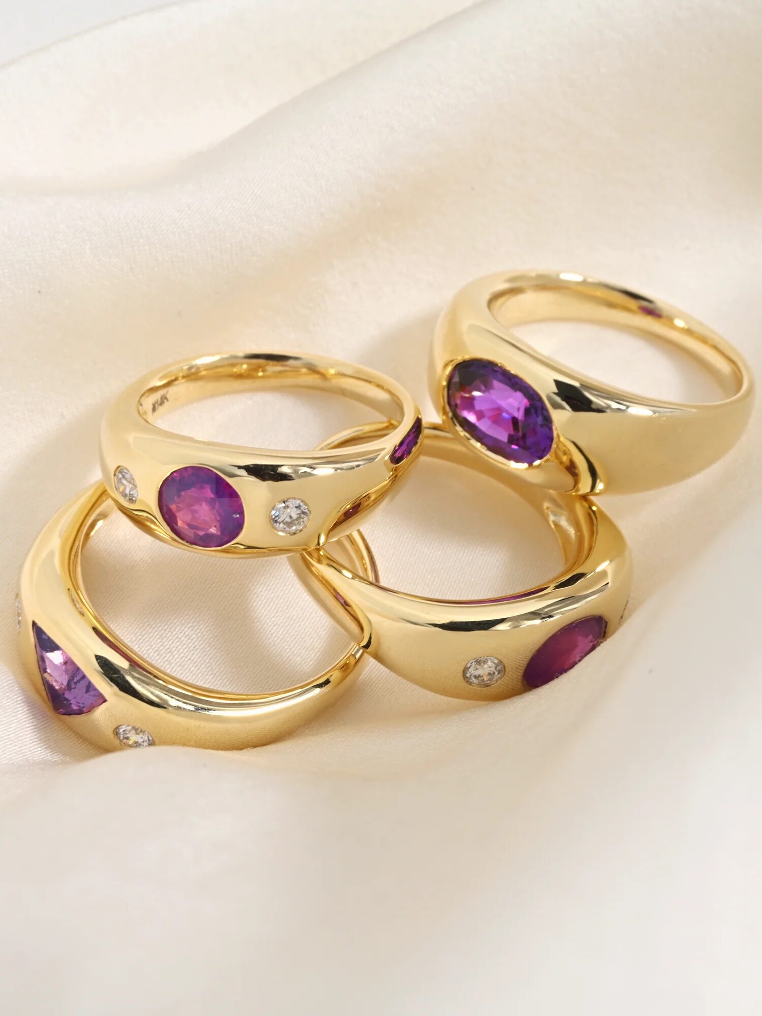 Four gold rings set with purple and white gemstones are placed on a white fabric background.