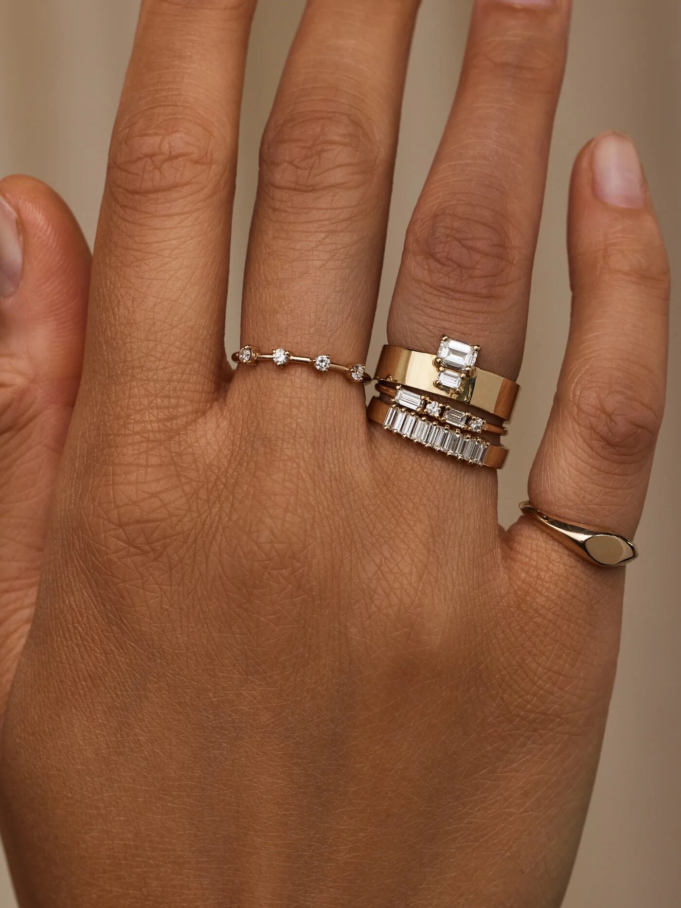 A hand wearing four different gold rings: a thin band with small stones, a wide band with a rectangular stone, a band with multiple small rectangular stones, and a simple thin band.