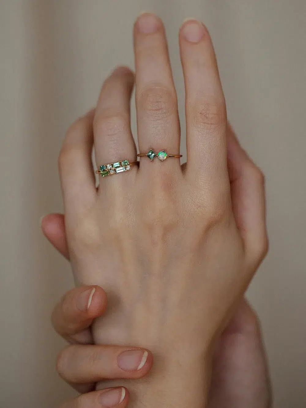 Two hands are clasped together, one with two rings on the ring and pinky fingers, both featuring green gemstones.