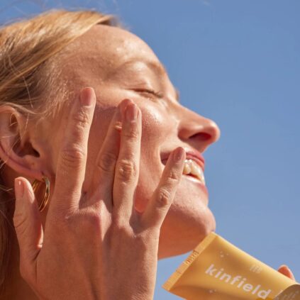 A person is applying sunscreen to their face while smiling under a clear blue sky. The sunscreen tube in their hand is labeled "kinfield.