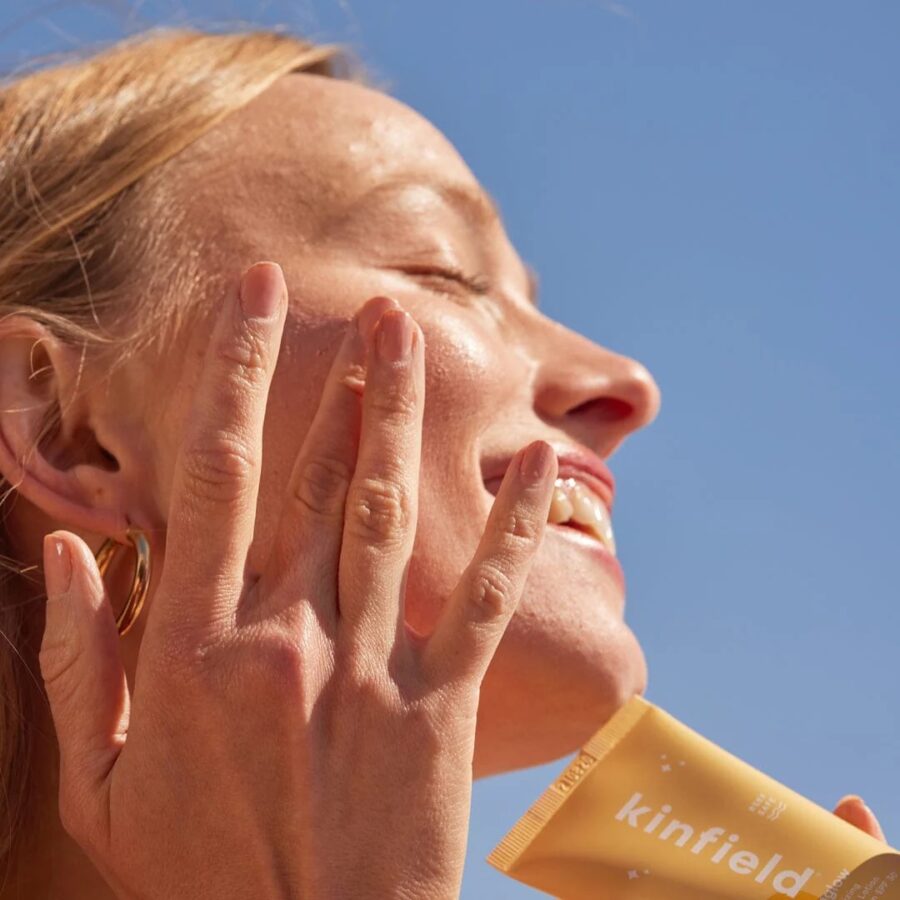 A person is applying sunscreen to their face while smiling under a clear blue sky. The sunscreen tube in their hand is labeled "kinfield.