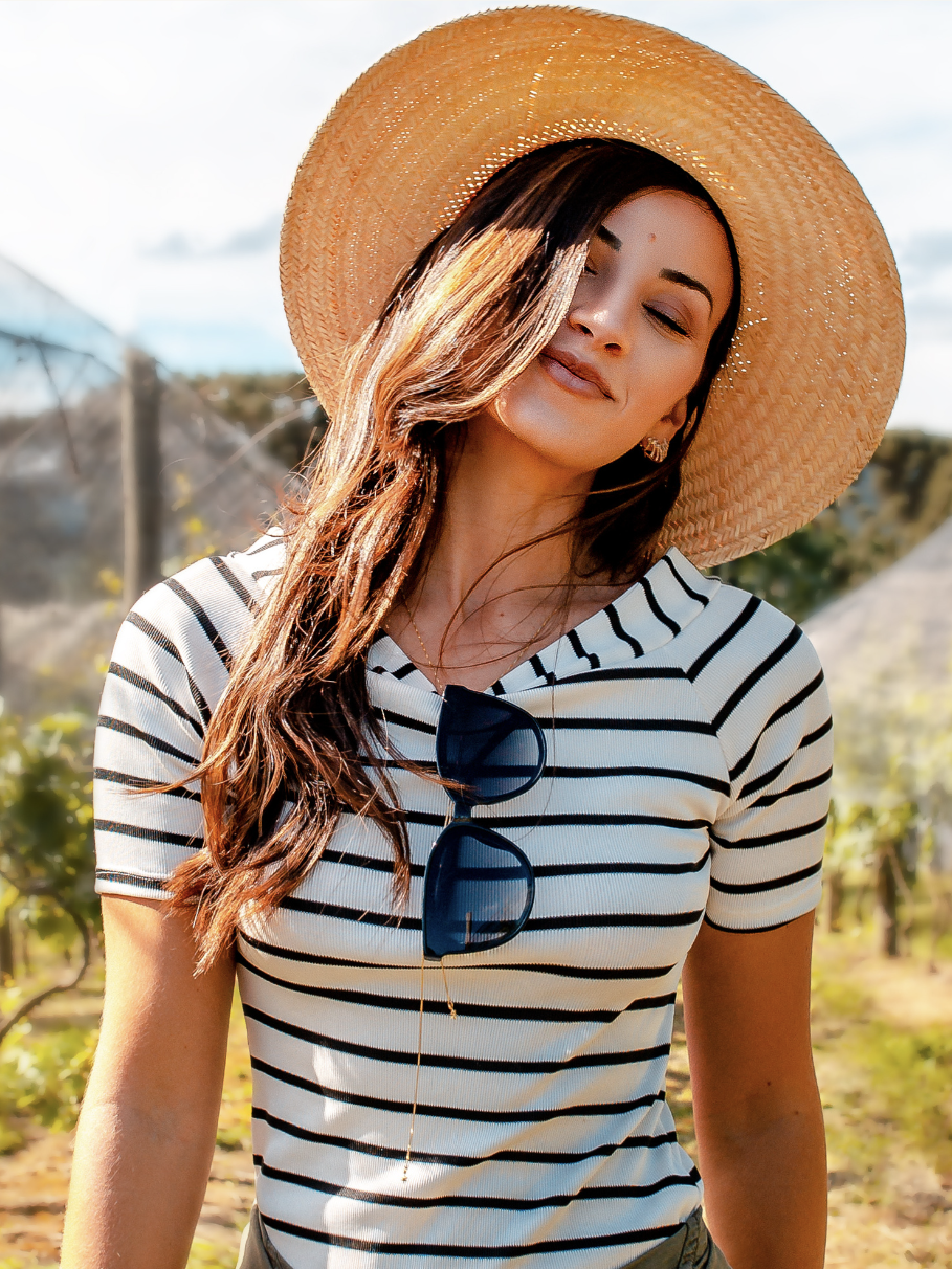 A woman in a straw hat and striped top stands in a vineyard, enjoying the sun, with sunglasses hanging from her shirt.