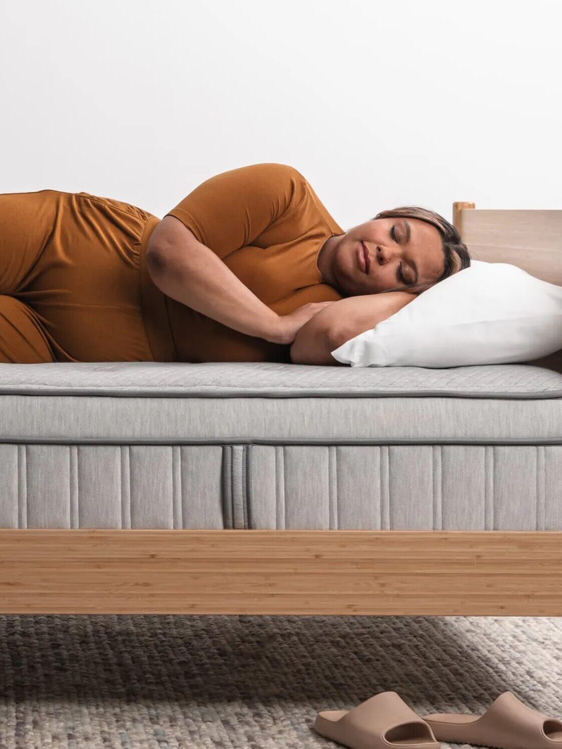 A person in an orange outfit lies on their side in a bed with a gray mattress, head resting on a white pillow. Tan slippers are placed on the floor nearby.