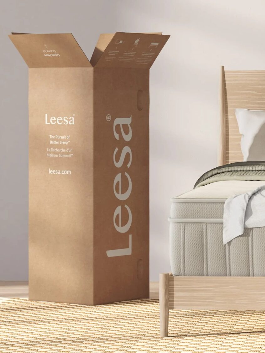 A Leesa mattress box stands open next to a partially visible bed in a well-lit room with wooden furniture and a textured rug.