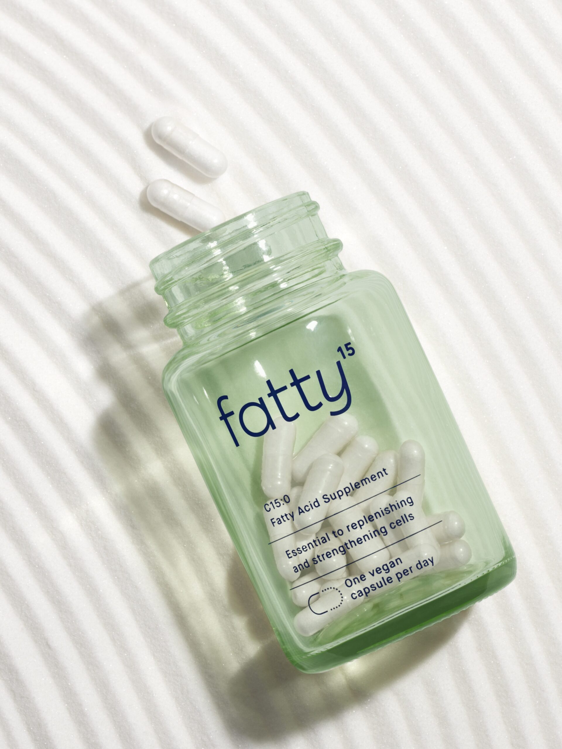 A green bottle labeled "fatty15" with white capsules spilling out. The label mentions it is a fatty acid supplement with one vegan capsule per day.