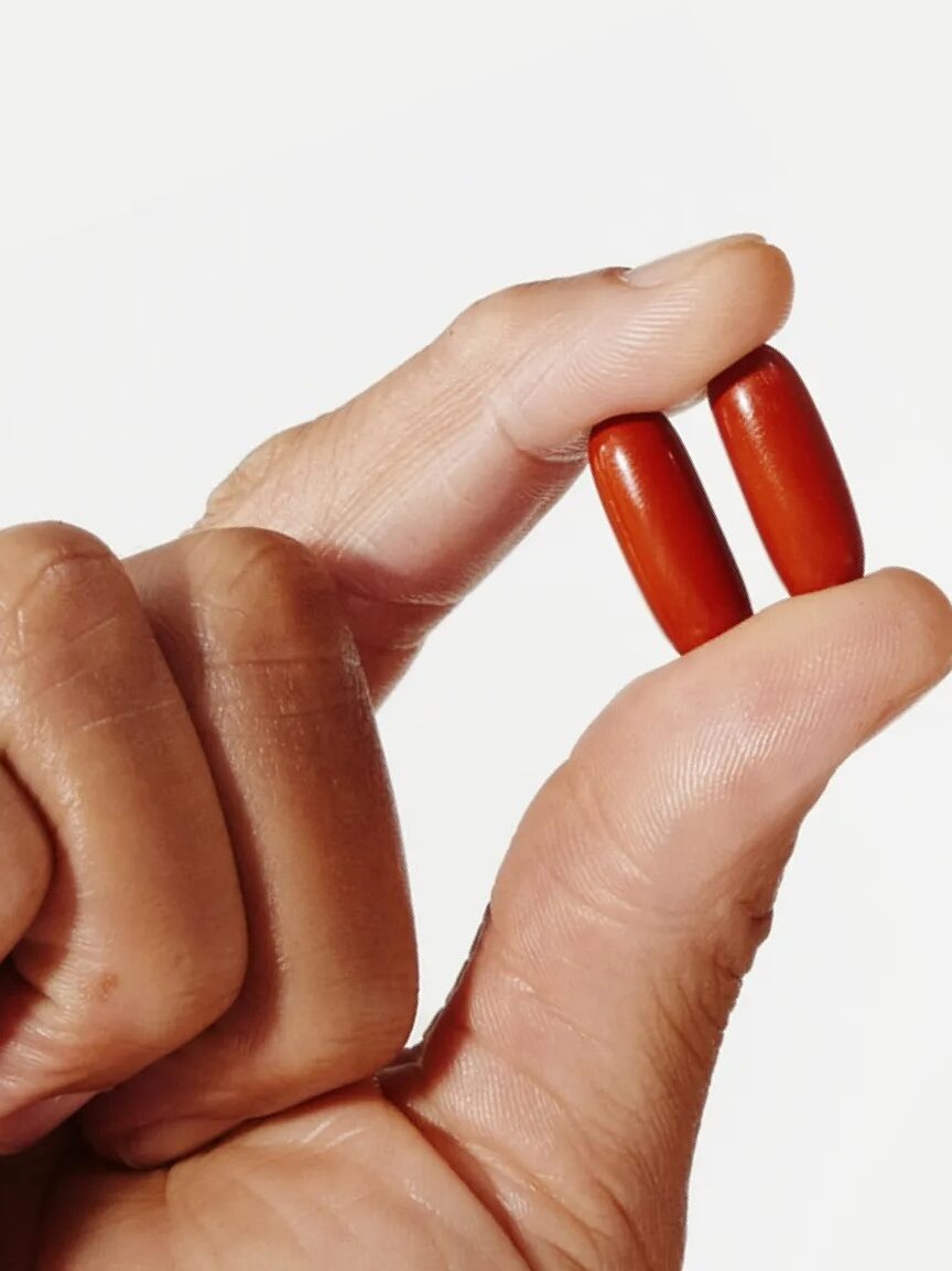 A hand holding two red pills between the thumb and index finger against a white background.