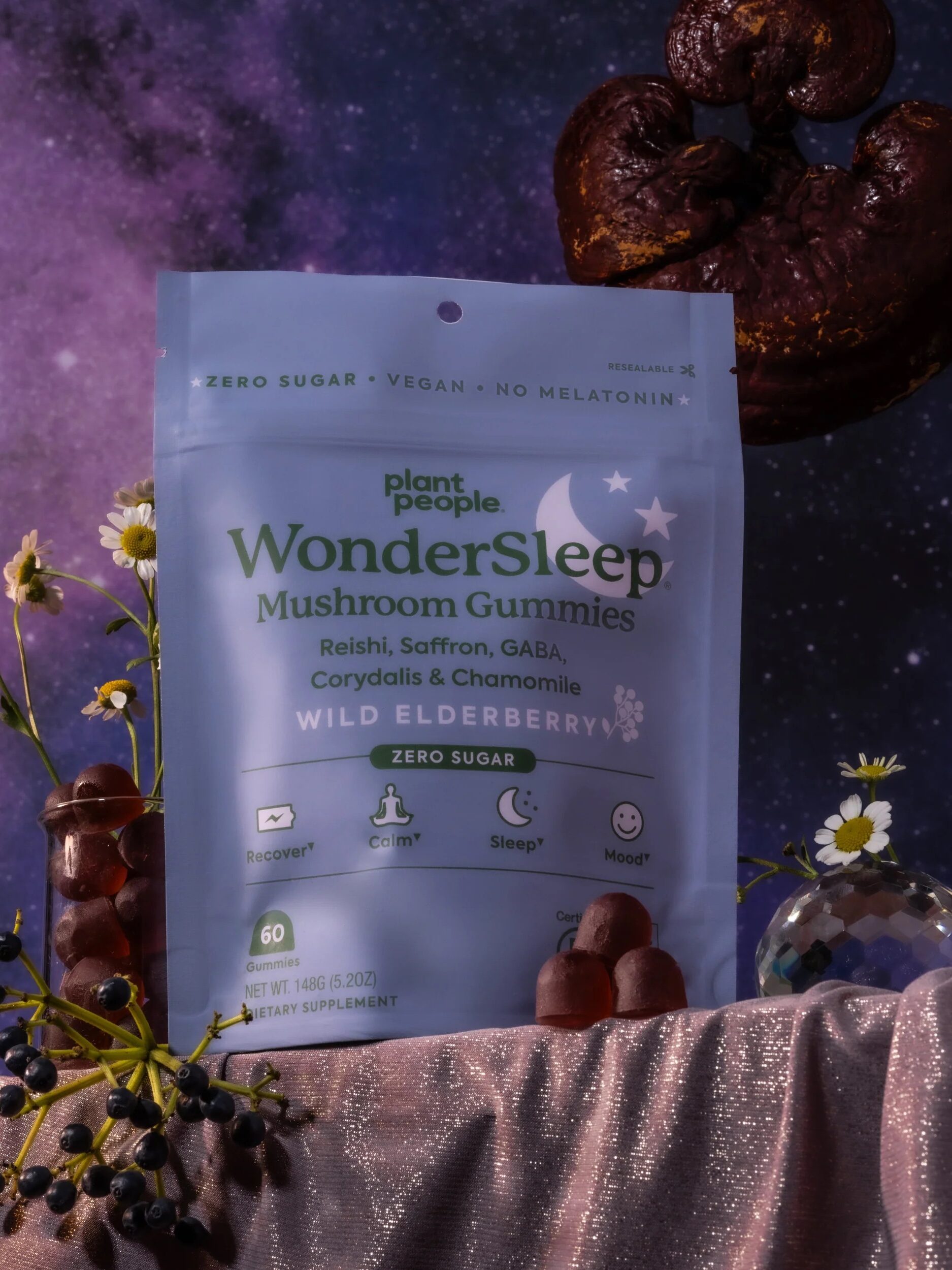 Package of Plant People WonderSleep Mushroom Gummies on a draped fabric surface, with two gummy bears, flowers, and a decorative mushroom. Starry night sky in the background.