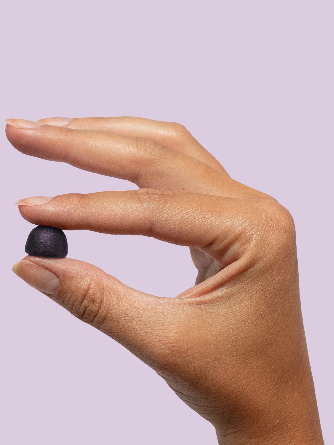 A hand holding a small, black, round object between the thumb and index finger against a plain, light purple background.