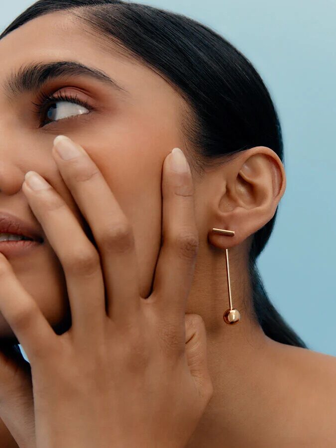 A woman touches her face, showcasing a long gold earring against a light blue background.