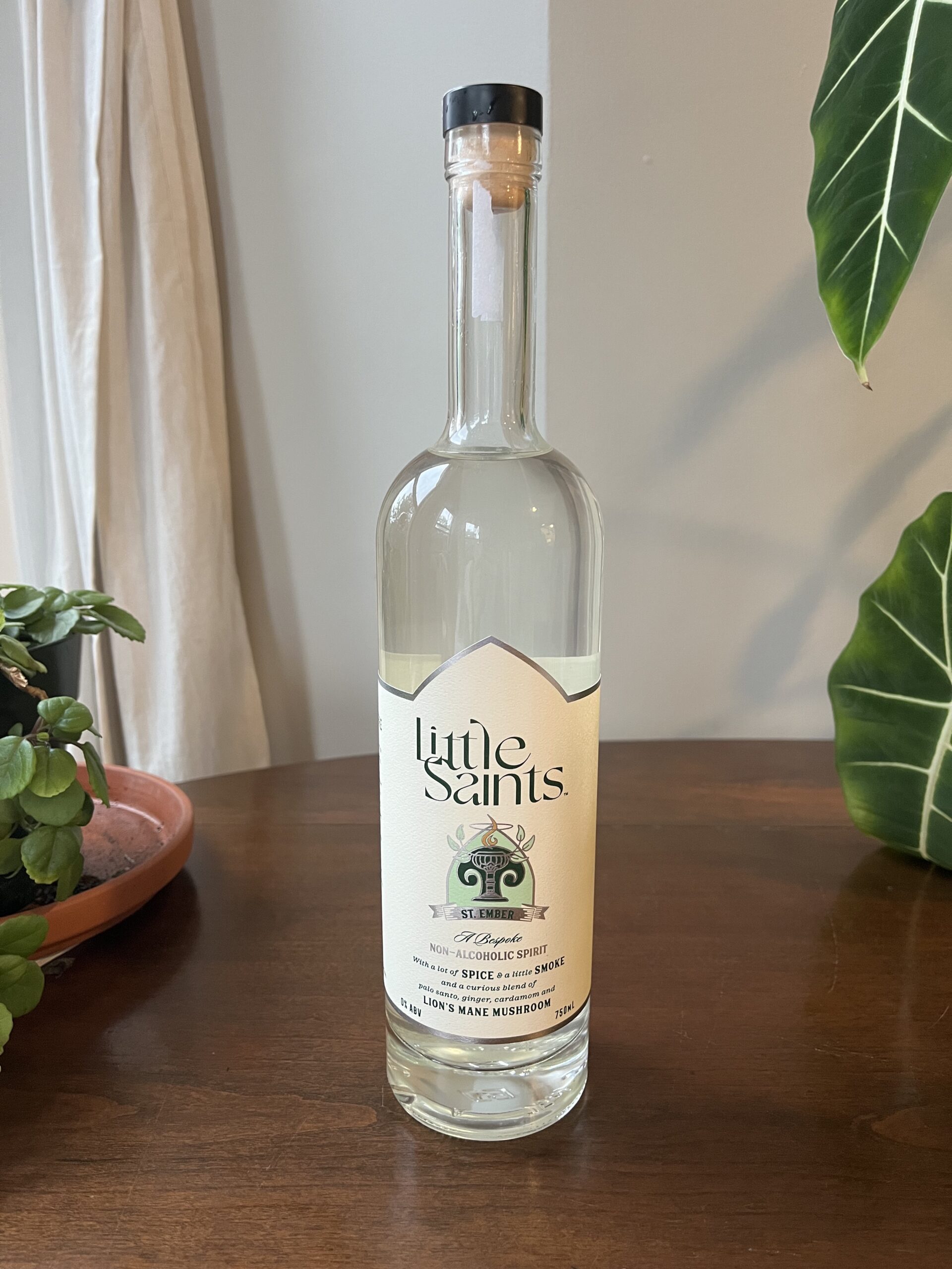 A clear glass bottle of Little Saints non-alcoholic spirit with adaptogens, placed on a wooden surface next to green potted plants.