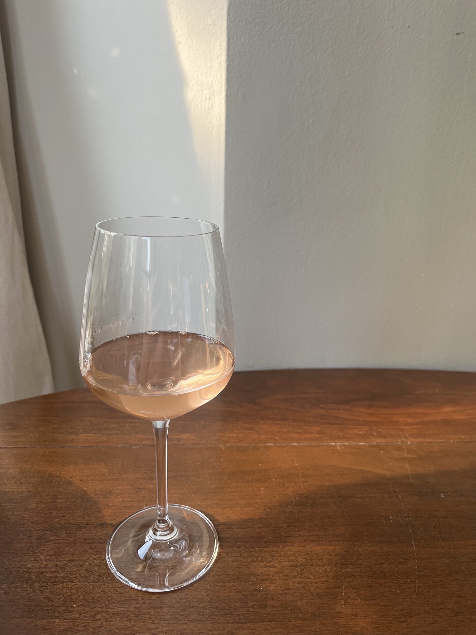 A half-full glass of rosé wine sits on a wooden table with soft natural light illuminating the scene.