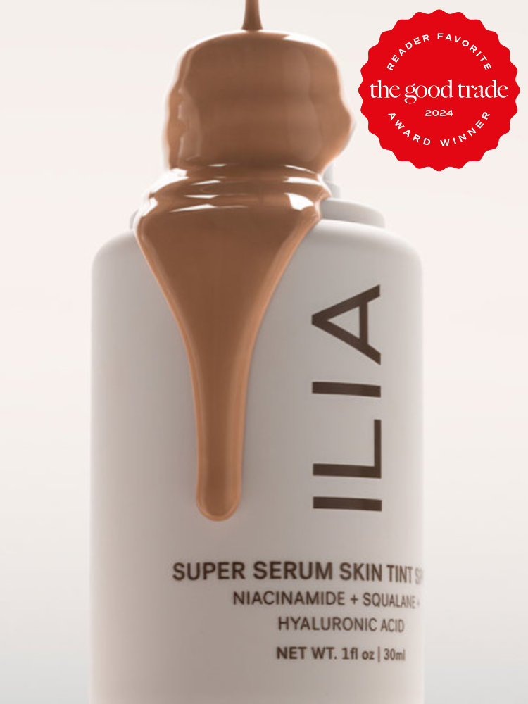 A white bottle of ILIA Super Serum Skin Tint with brown liquid oozing over the top. The bottle is labeled with ingredients like niacinamide, squalane, and hyaluronic acid. A red award badge is in the corner.