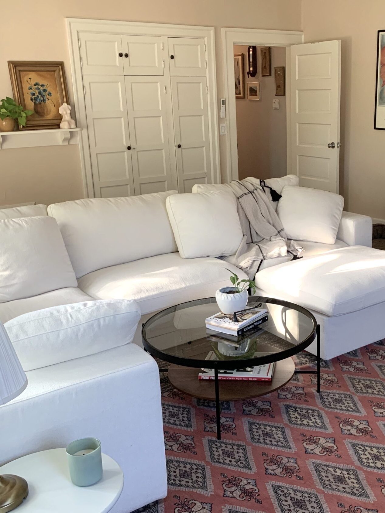 A Good Trade Editor's living room with a white sectional sofa from 7th Avenue, a glass coffee table, a brown armchair, and a red patterned rug. Artwork and shelves with decor are on the walls. A dog bed is on the floor.