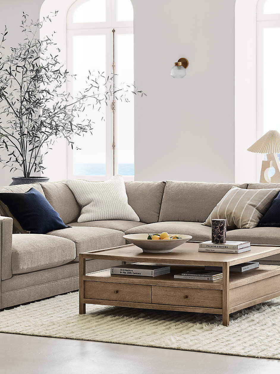 A modern living room with a beige sectional sofa, wooden coffee table, white rug, and large windows overlooking a coastal landscape. A bowl of fruit and decorative items are on the coffee table.