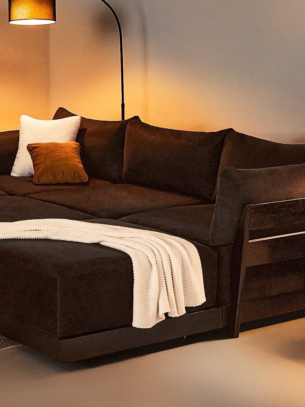 A cozy living room with a dark brown sectional sofa, a white blanket, a round wooden coffee table, a floor lamp, a side table with a lamp, shelves, a fireplace, and a potted plant.