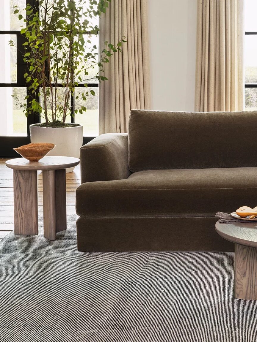 A living room with a large brown sofa, two side tables, and a coffee table. The coffee table holds a bowl, books, and a glass of water. Large windows and beige curtains are in the background.