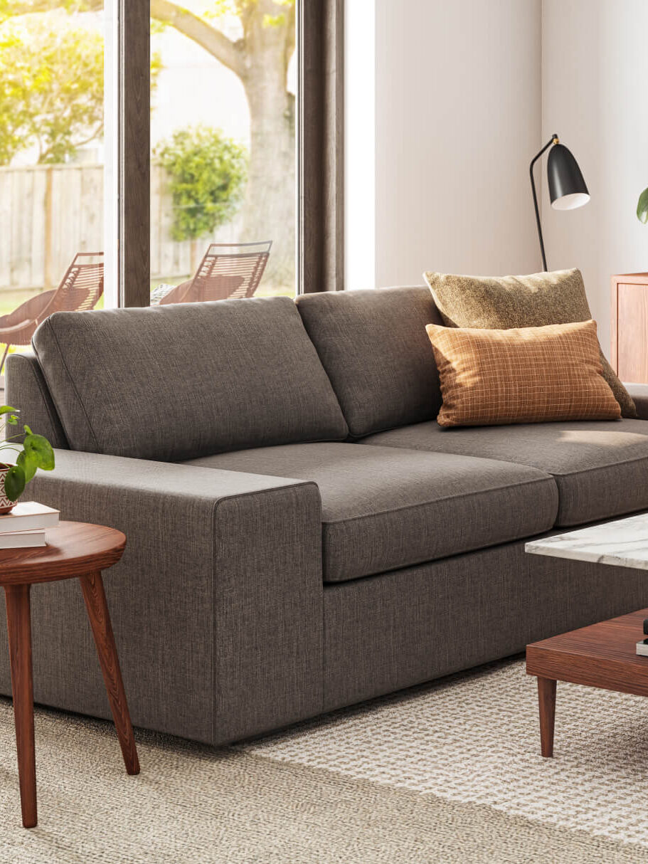 A modern living room with a grey sofa, a wooden side table with a plant, a marble coffee table, and a wooden credenza with plants and decor. Large windows show an outdoor patio.