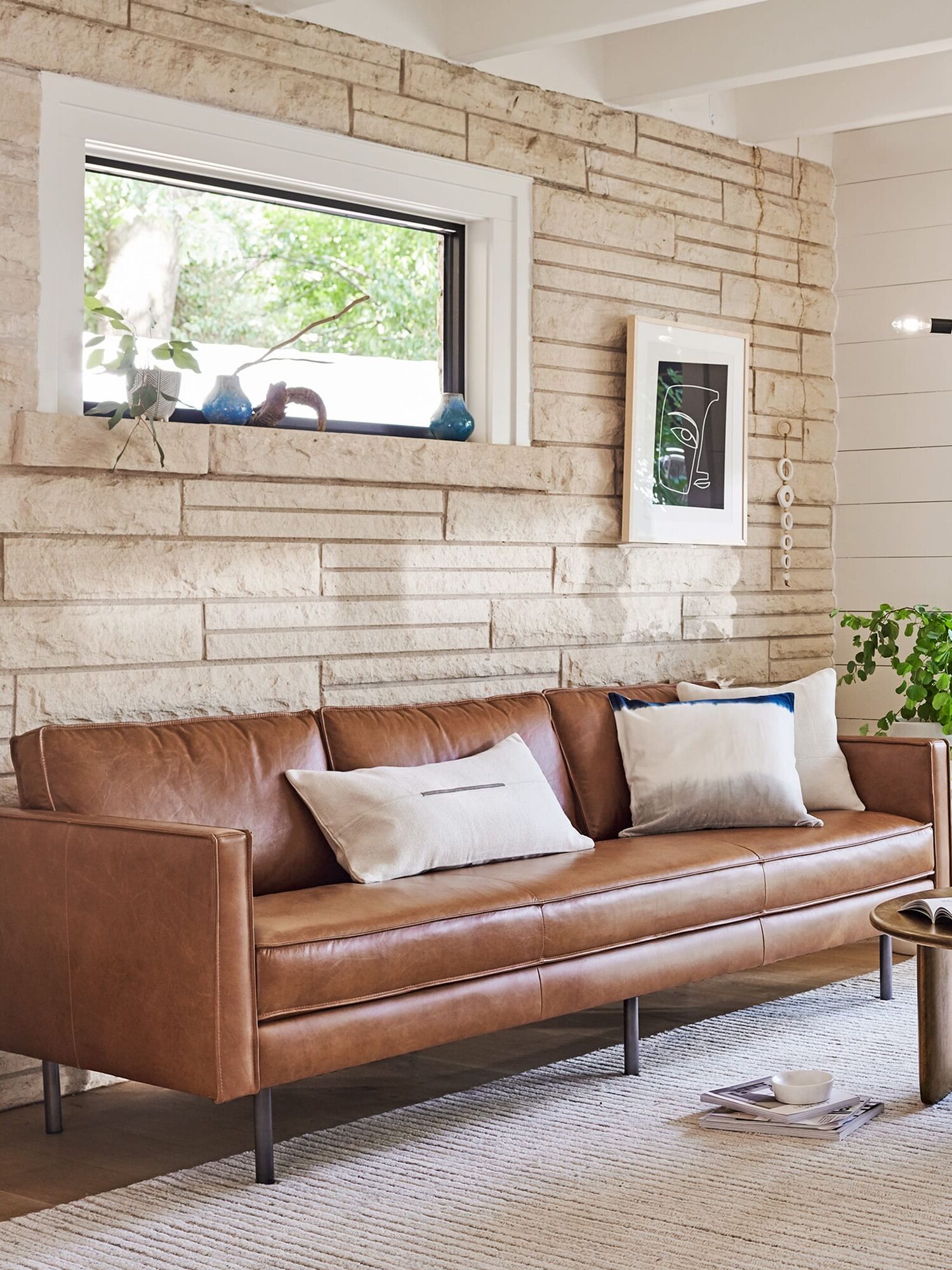 Living room with a tan leather sofa, white and gray cushions, a round wooden coffee table, a plant, wall art, and a large window. Light fixture hanging from the ceiling and a neutral-colored rug on the floor.