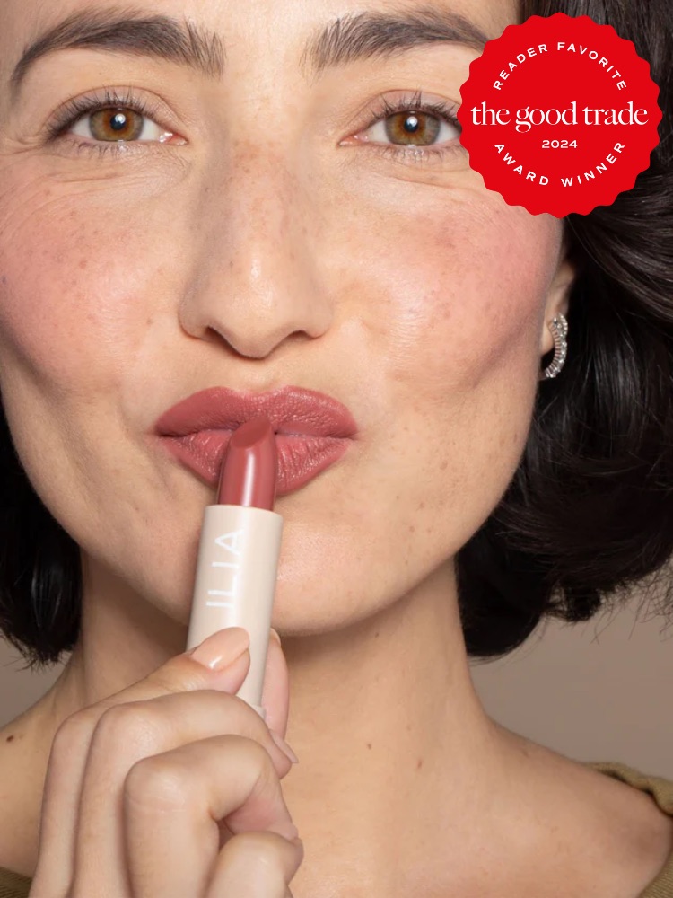 A person with short dark hair applies pink lipstick. A "Reader Favorite" award badge from "The Good Trade" for 2024 is displayed on the top right corner.