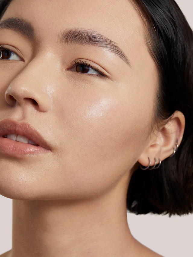 A close-up of a person with short dark hair, smooth skin, and silver hoop earrings looking slightly off to the side, against a neutral background.