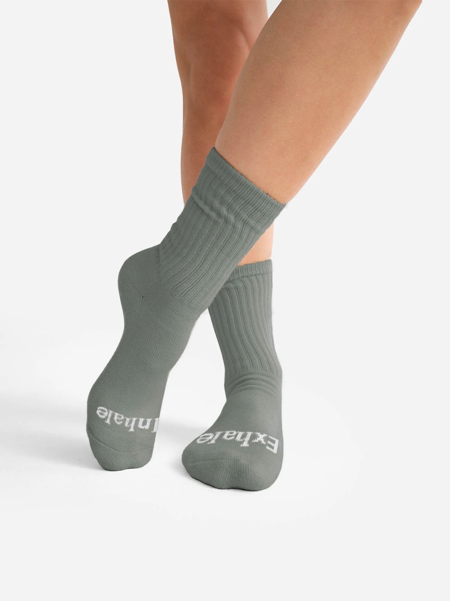 A person wearing gray ribbed organic socks with the word "Exhale" printed on the toes.