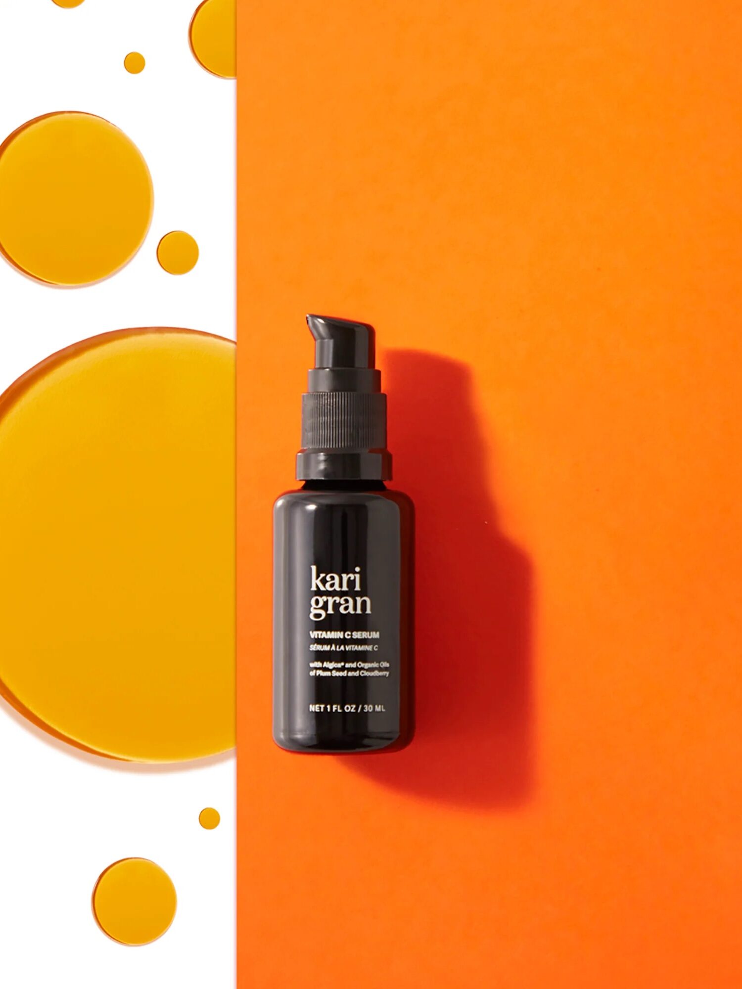 A black bottle of Kari Gran Vitamin C Serum is placed vertically on an orange and white background with scattered yellow oil droplets.