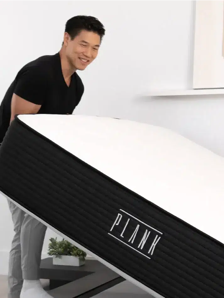 A person in a black shirt and gray pants is flipping a black and white mattress labeled "PLANK." An icon indicating the mattress is flippable is visible in the lower-left corner.