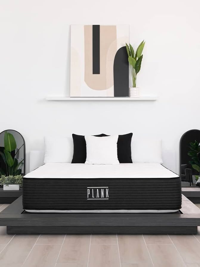 A minimalistic bedroom with a platform bed featuring a 'PLANK' mattress, black and white pillows, modern decor, and plants on side tables and a wall shelf.