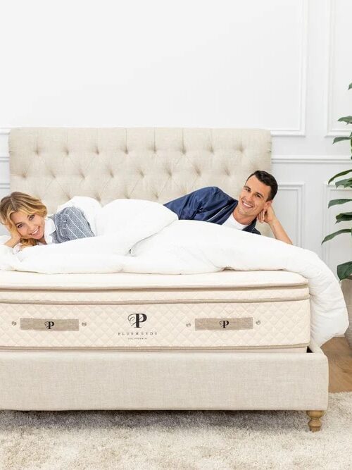 A couple lies on a bed with a beige tufted headboard, surrounded by white bedding. A nightstand with a lamp is on the left; a potted plant is on the right. The room has a light-colored rug and hardwood floor.