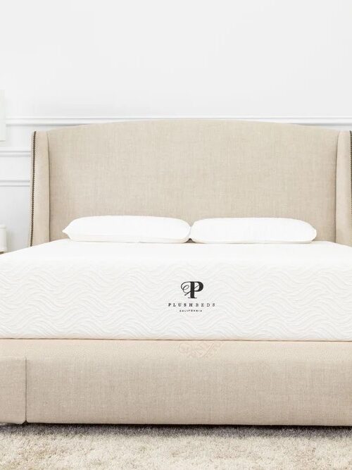 A beige upholstered bed with a mattress labeled "Plushbeds" stands between two white nightstands with gold accents and matching table lamps in a neatly arranged bedroom.