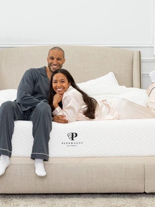 A smiling couple in pajamas sit and lay on a bed in a bright room with twin bedside tables and lamps.