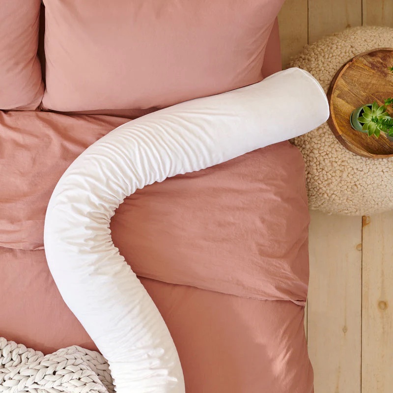 A white body pillow with a curved shape is placed on a pink bedspread next to a small round wooden table with a green plant.