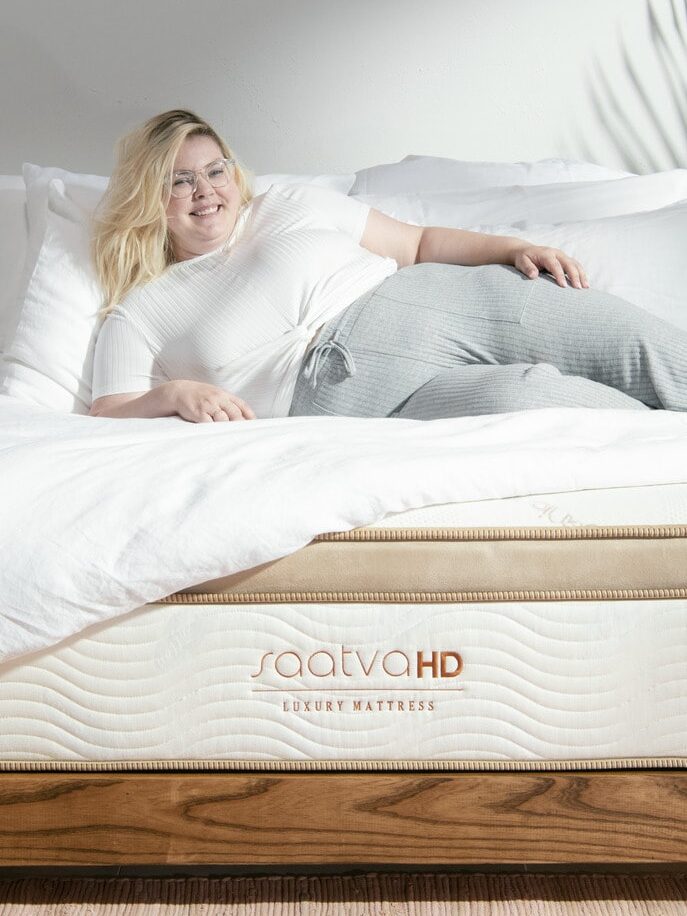 A person lying on a Saatva HD luxury mattress, dressed in a white shirt and gray pants, with a content expression.