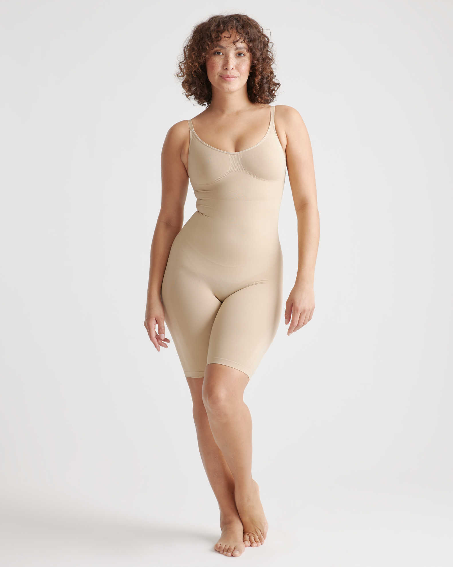 A person with curly hair is wearing a beige, form-fitting bodysuit. They are barefoot and standing against a plain white background.