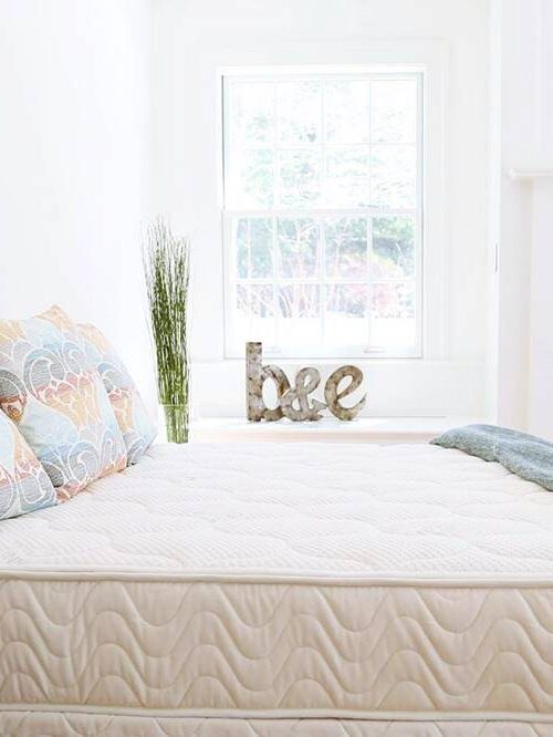 A neatly made bed with patterned pillows, a light blue throw, and a glass of water on a side table in a bright bedroom with a fireplace and decor accent spelling "bee".