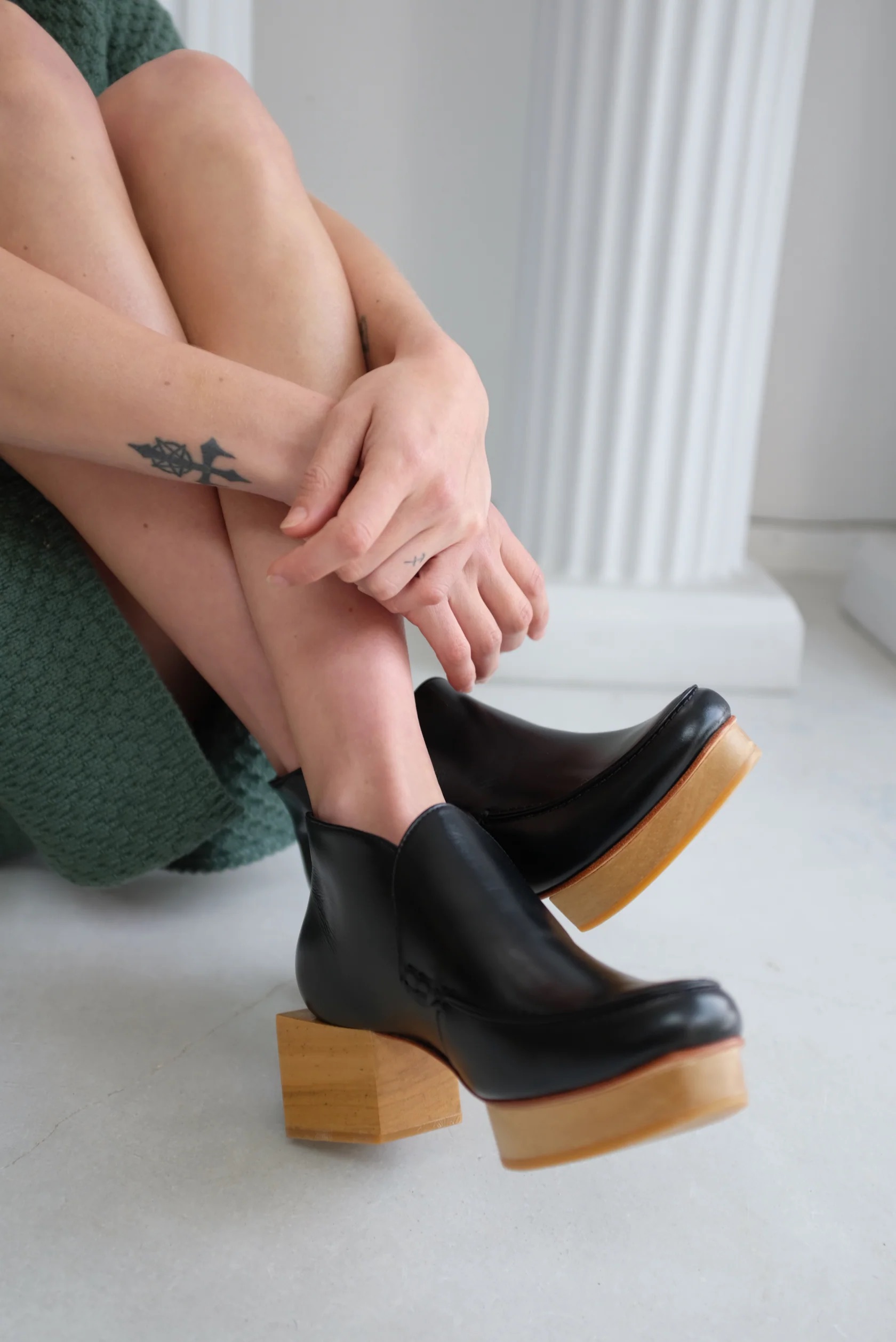 Person with a star tattoo on their wrist sits with legs crossed, wearing black leather boots with wooden heels.