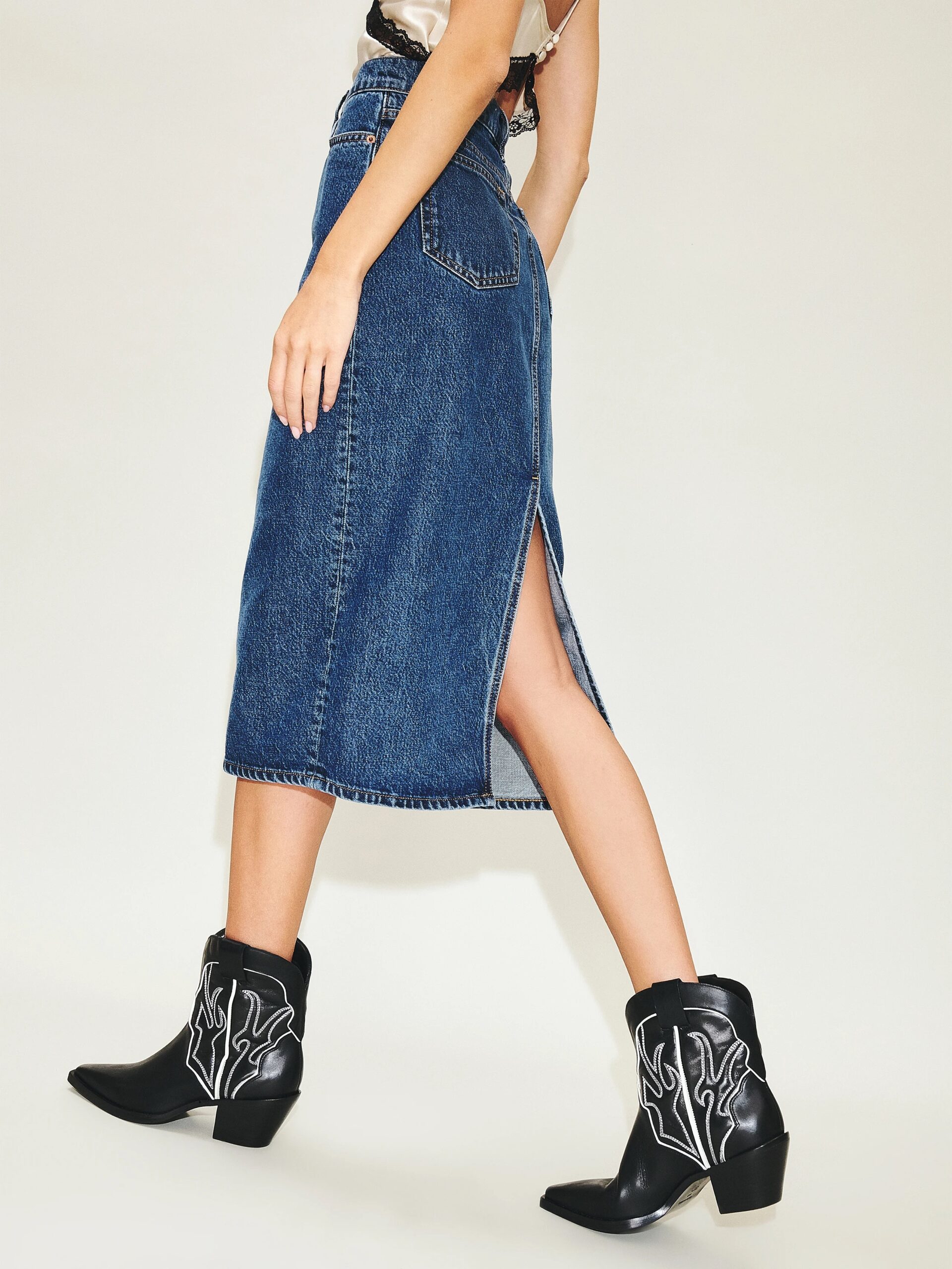 Person wearing a denim midi skirt with a side slit, paired with black cowboy boots featuring white flame detailing, walking against a plain background.