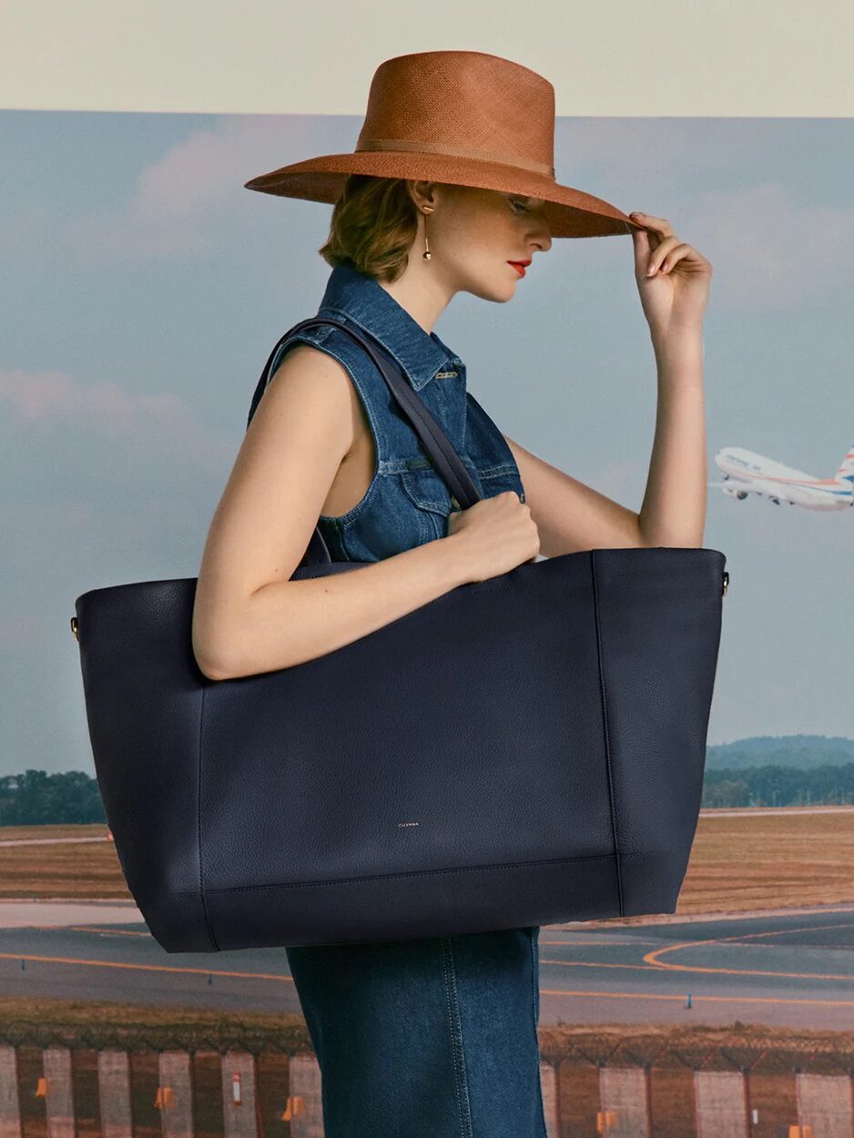 A woman in a wide-brimmed hat and denim vest carries a large tote bag, touching her hat, with an airplane and runway in the background.