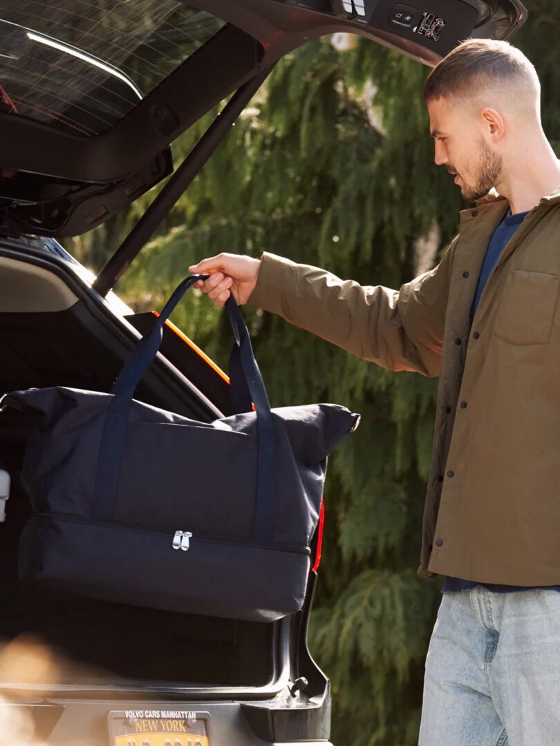 Man loading a large blue bag into the trunk of a car in a wooded outdoor area.