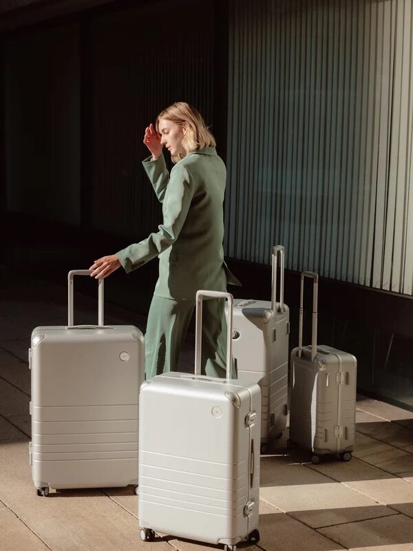 A woman in a green suit stands beside three white suitcases, adjusting her hair in a sunlit, modern corridor.